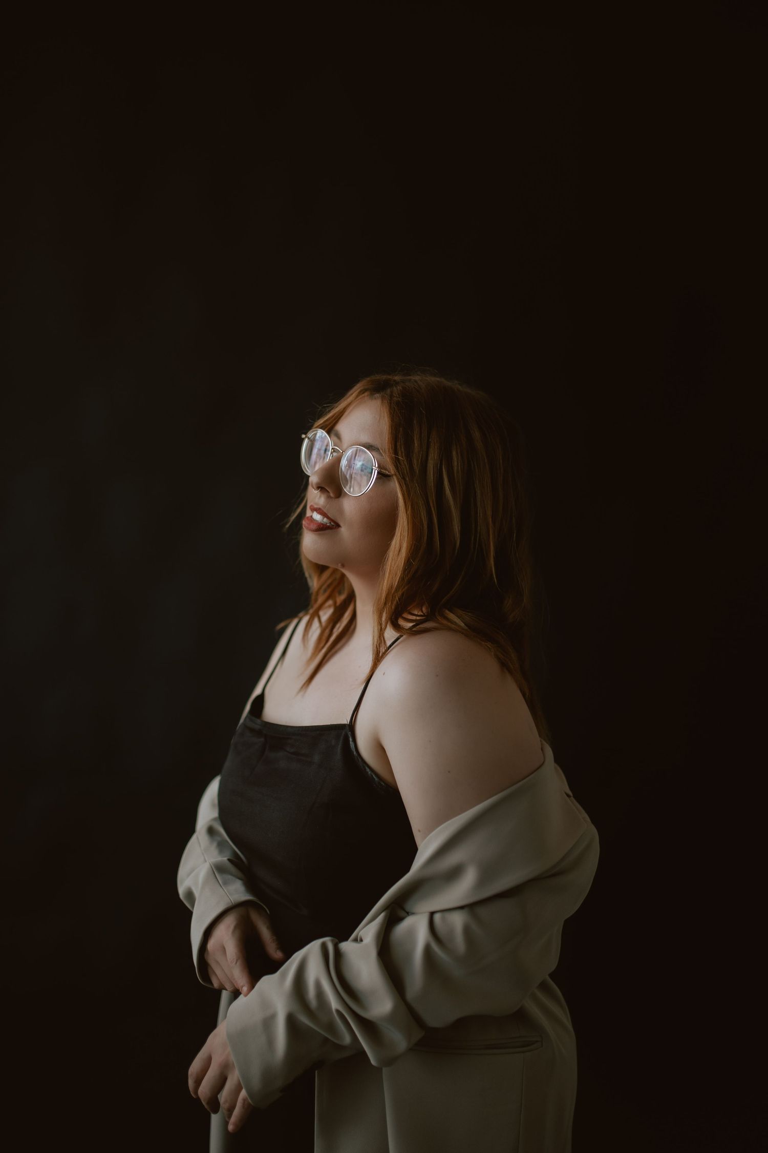 A woman in glasses and an oversized jacket poses confidently against a dark background.