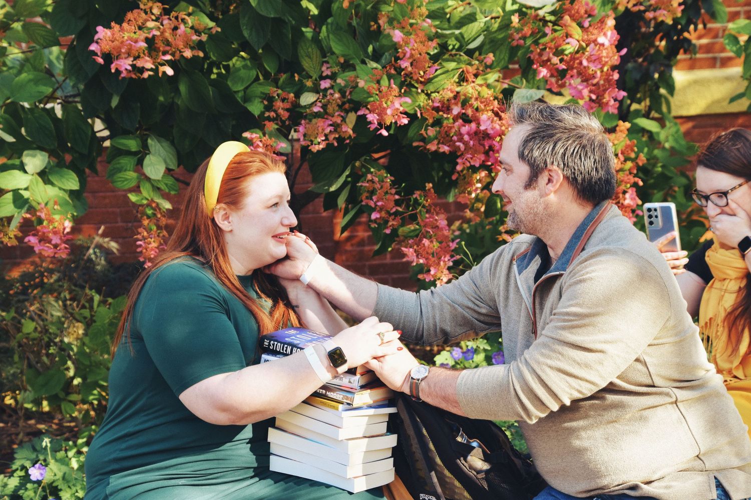 A man tenderly touches a red-haired woman's face as they share a joyful moment outdoors among pink flowers.