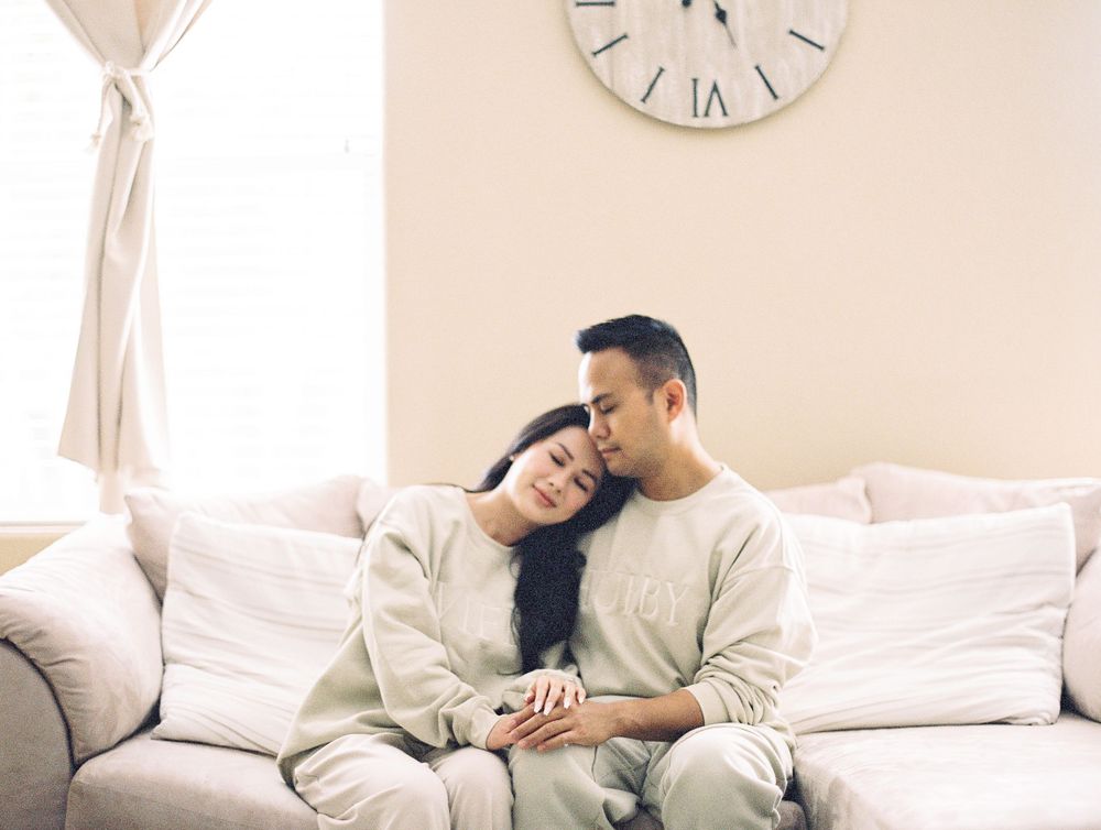 Couple cuddling on light colored sofa beneath wall clock in matching casual outfits.