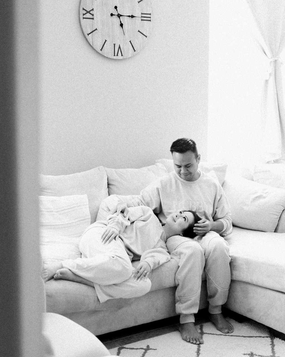 Couple in matching loungewear relaxing on sectional sofa under wall clock in black and white.