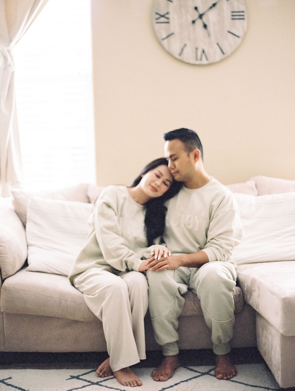 Couple relaxing together on gray sectional sofa under wall clock in matching beige outfits.