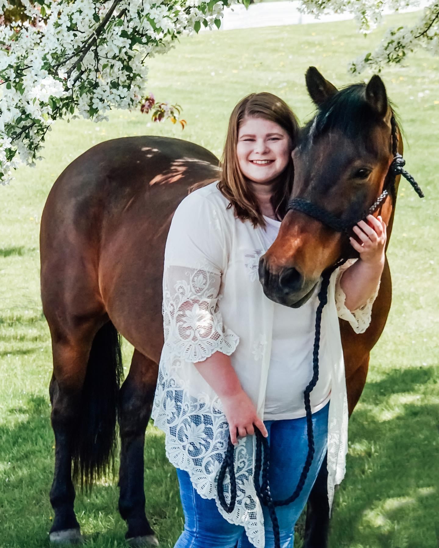 A beautiful bay horse poses with someone wearing a white lace top in a sunny field.