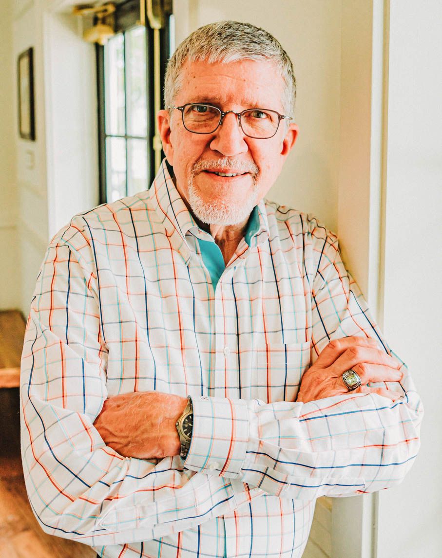 A senior wearing glasses and a plaid button-down shirt stands with arms crossed in a casual indoor setting.