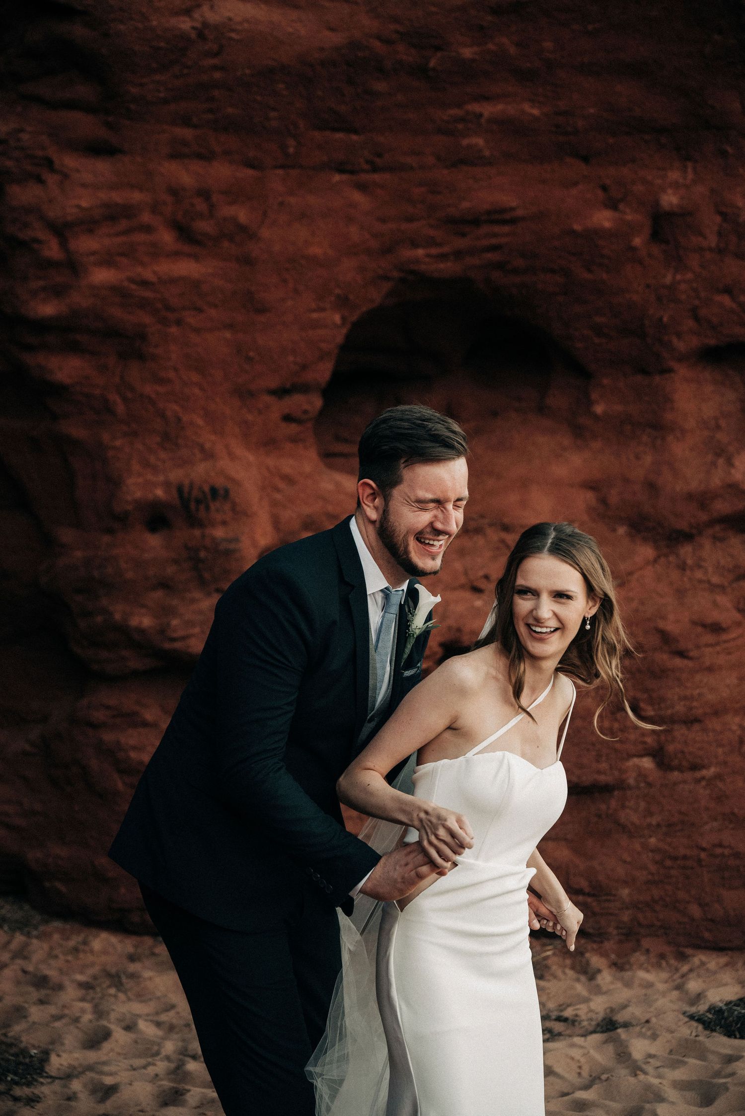 Joyful wedding couple sharing a laugh against red rock formation.