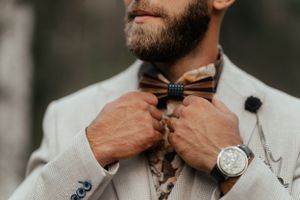 A stylish man with a beard and a watch carefully adjusts his dapper bow tie.