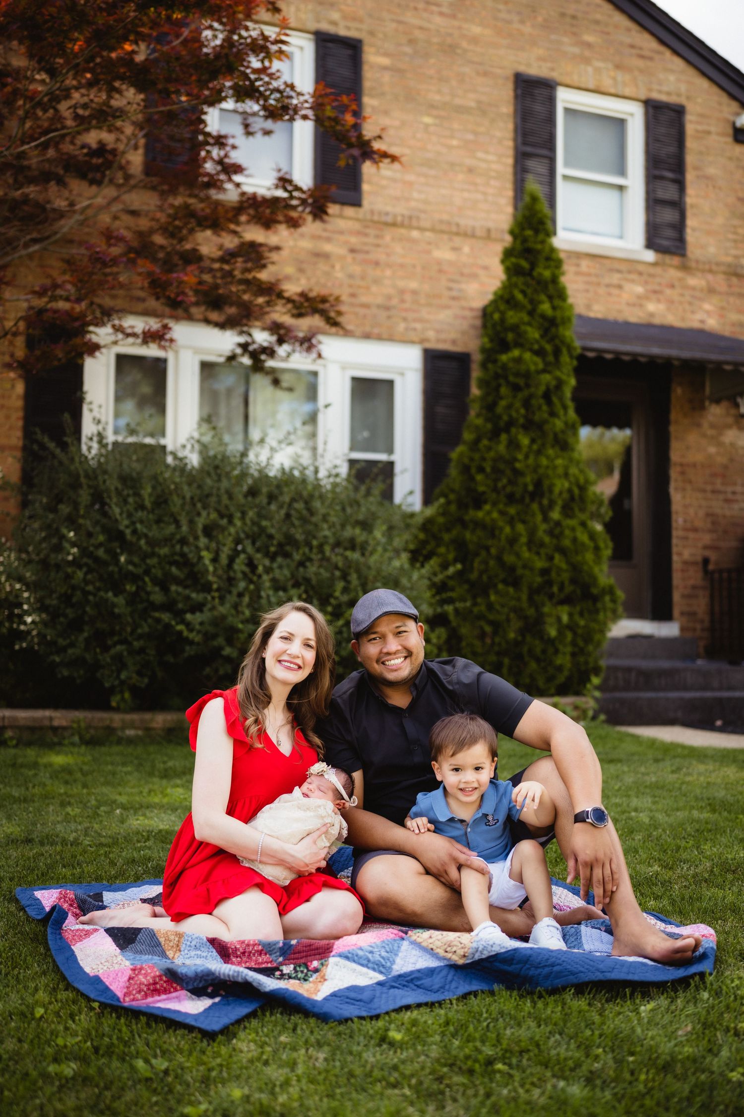 A family sits on a patchwork blanket for a portrait in front of their brick house.