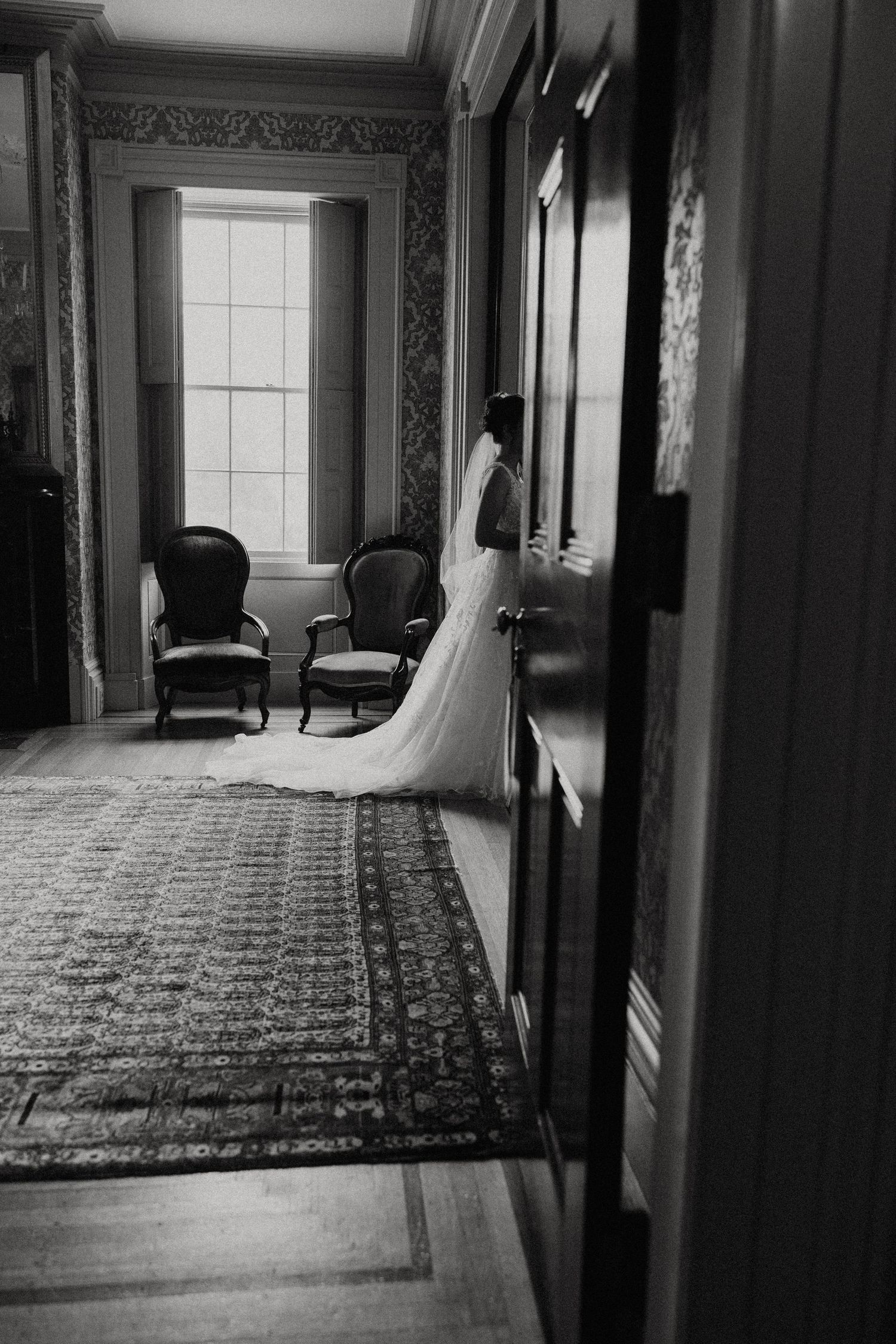 Black and white photo of bride in wedding gown standing by window in elegant vintage room. New Bedford, Ma Miranda Photo & Co