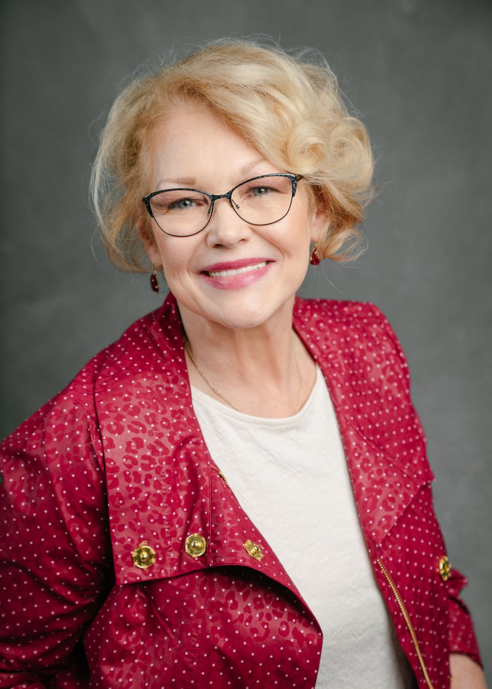 Smiling woman in red patterned jacket and glasses against grey background.