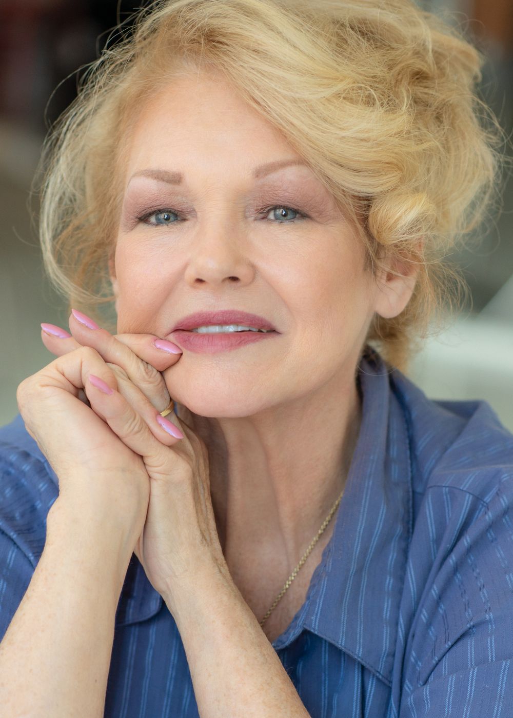 Portrait of an elegant woman in a blue shirt with blonde hair resting her chin on her clasped hands.