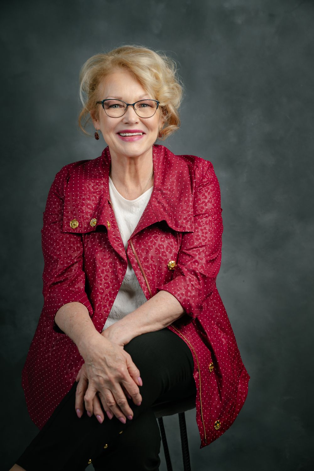 Stylish woman in a red patterned jacket and glasses seated against a dark background.