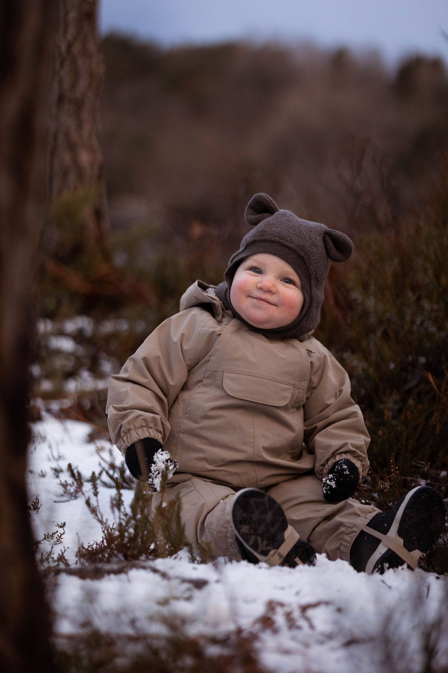Small figure in tan snowsuit and bear hat sits in snowy winter landscape.
