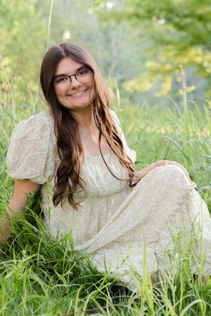 Someone wearing glasses and a cream colored dress sits in tall summer grass with a bright natural background.