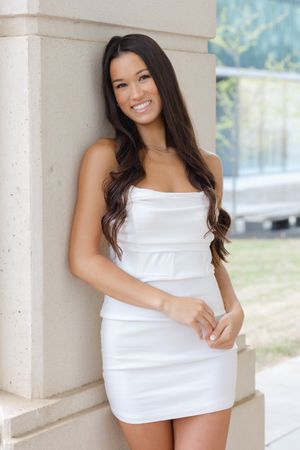 Woman in a white strapless dress poses against a pillar backdrop while beaming with a bright smile.