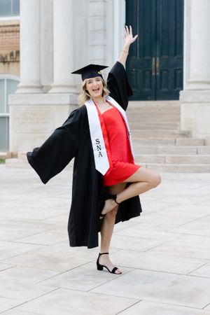 Graduate in cap and gown celebrates while wearing red dress and posing on marble steps.