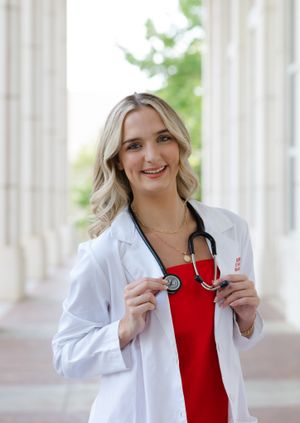 Healthcare professional in white lab coat and red top poses with stethoscope near columns in outdoor setting.