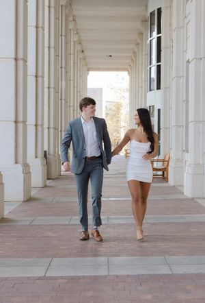 A couple walks hand in hand along an elegant columned walkway, with him in a suit and her in a white dress.