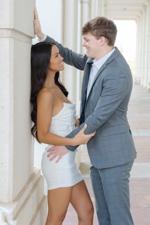 A couple embraces intimately in an elegant hallway, with her in a white dress and him wearing a grey suit.