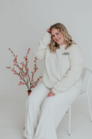 Person in cozy white sweatsuit lounges in chair next to decorative flowering branches against light backdrop.