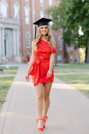 Graduate posing on campus path wearing red wrap dress, graduation cap, and matching red heels.
