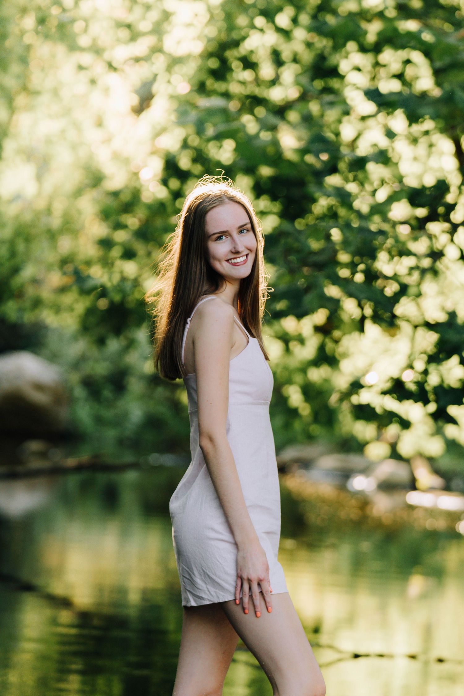 Someone in a white dress poses next to a pond on a sunny summer day with bokeh lighting.