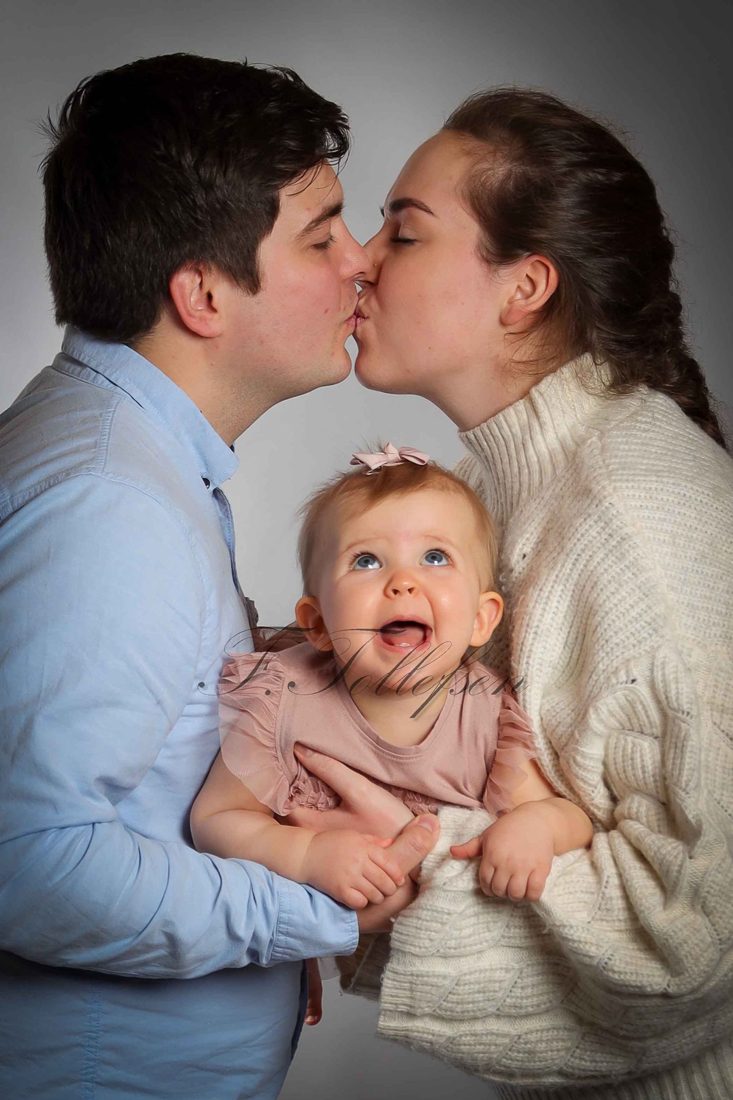 Young couple kissing while holding their wide-eyed baby girl in a studio portrait.
