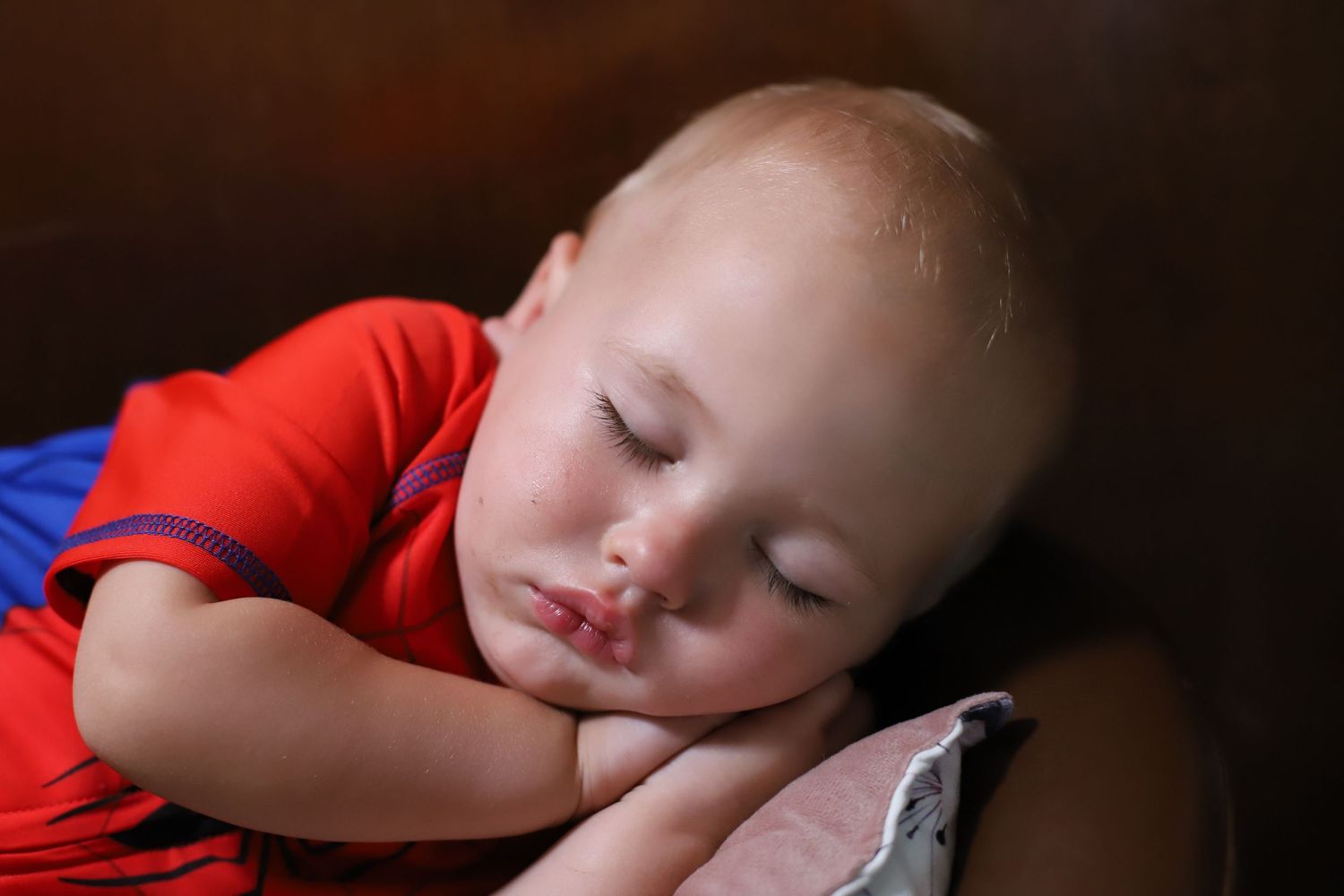 Adorable toddler boy in a red shirt sleeping peacefully with hands under his cheek on a cushion.