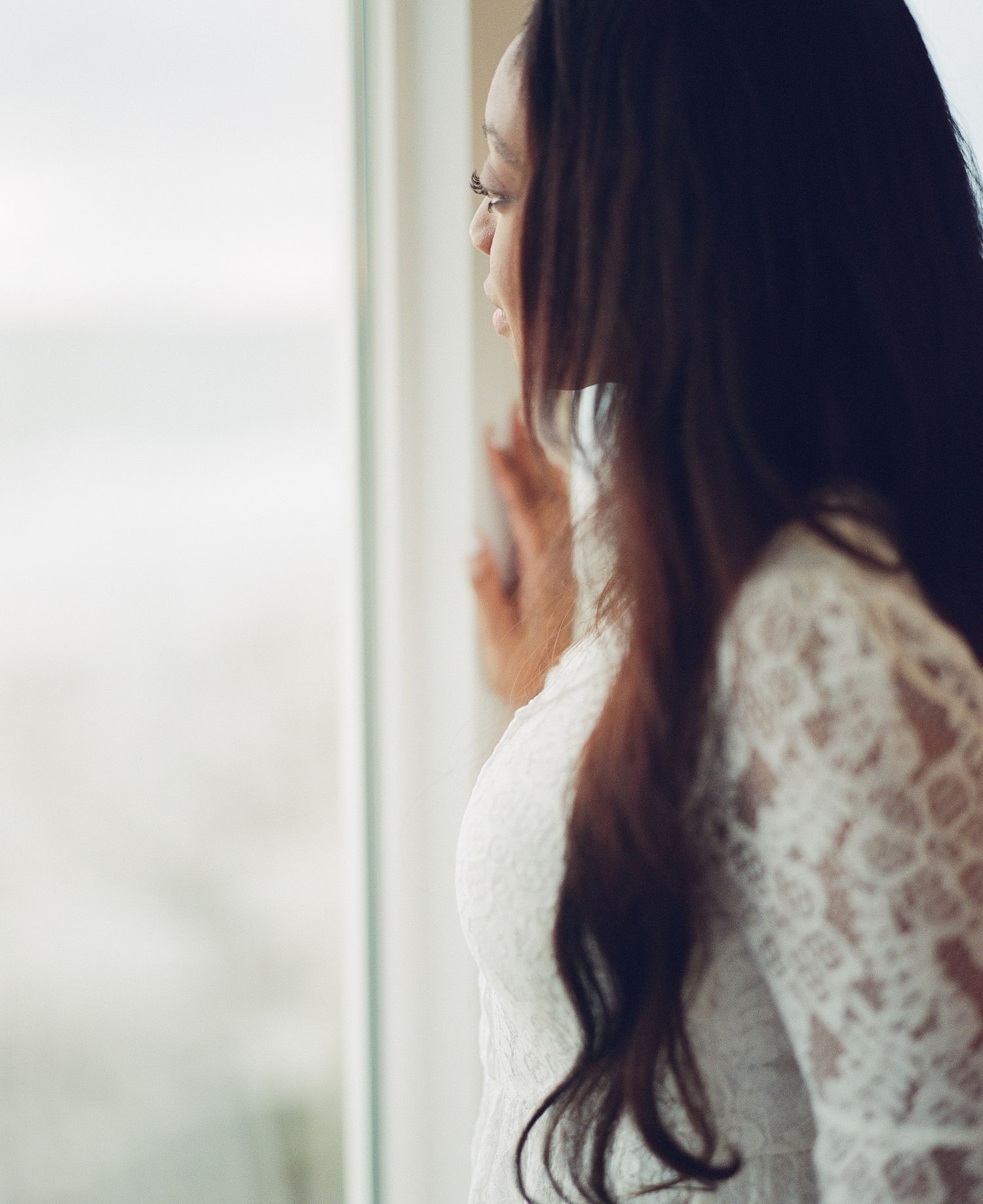 Woman in white lace dress gazing through window in soft dreamy lighting.