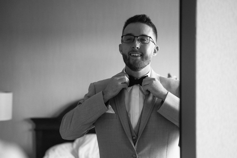 A groom adjusts his bowtie while getting ready in formal attire.