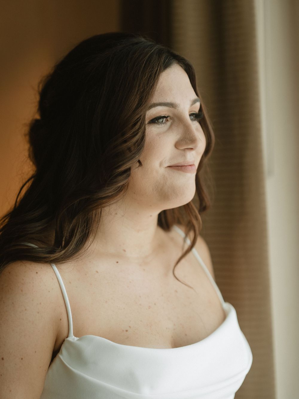 A bride in a white dress looks off to the side with natural window lighting during wedding preparations.