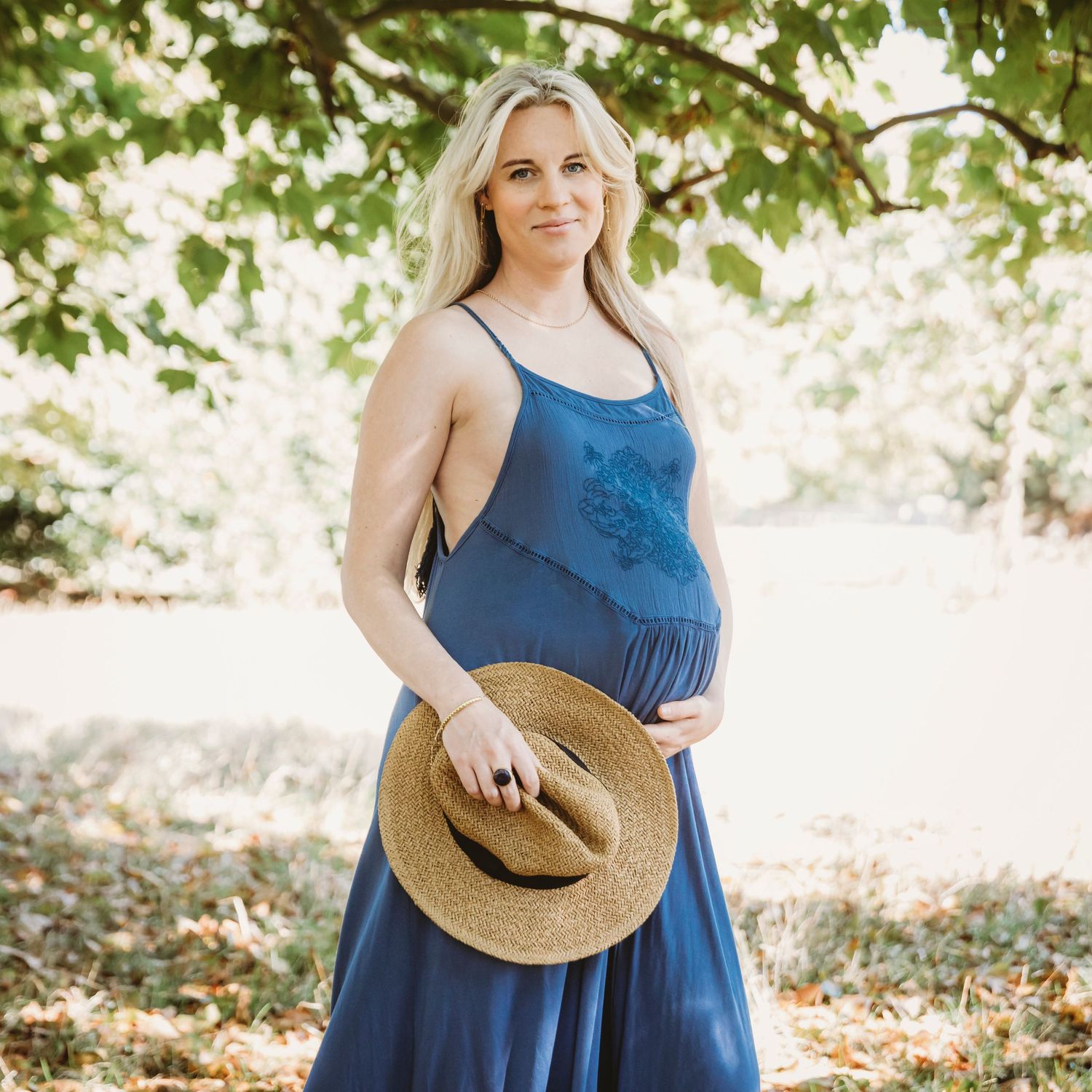 Maternity portrait in blue dress holding straw hat under tree canopy.