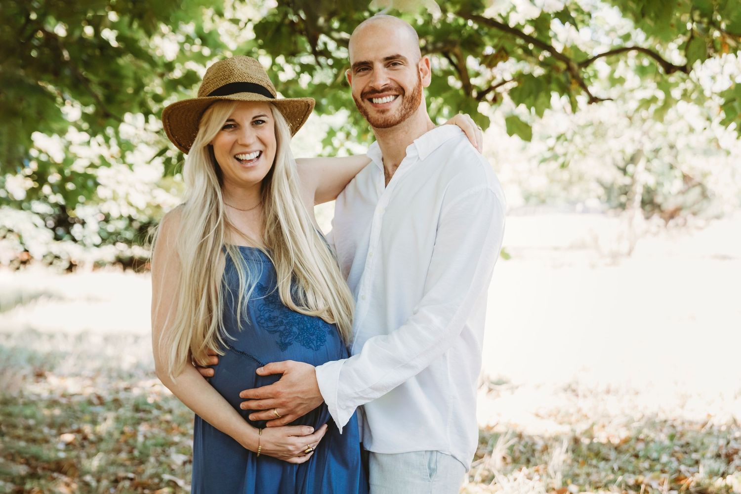 A couple poses outdoors in casual attire during a maternity photoshoot under green trees.