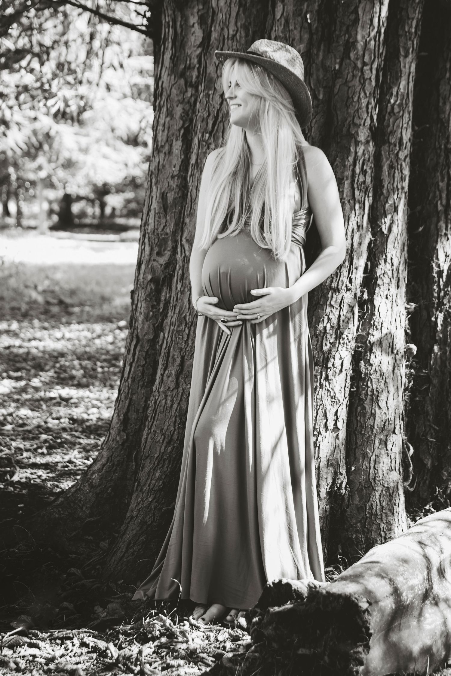 A maternity photo of a woman in a long maxi dress standing beside a large tree trunk.