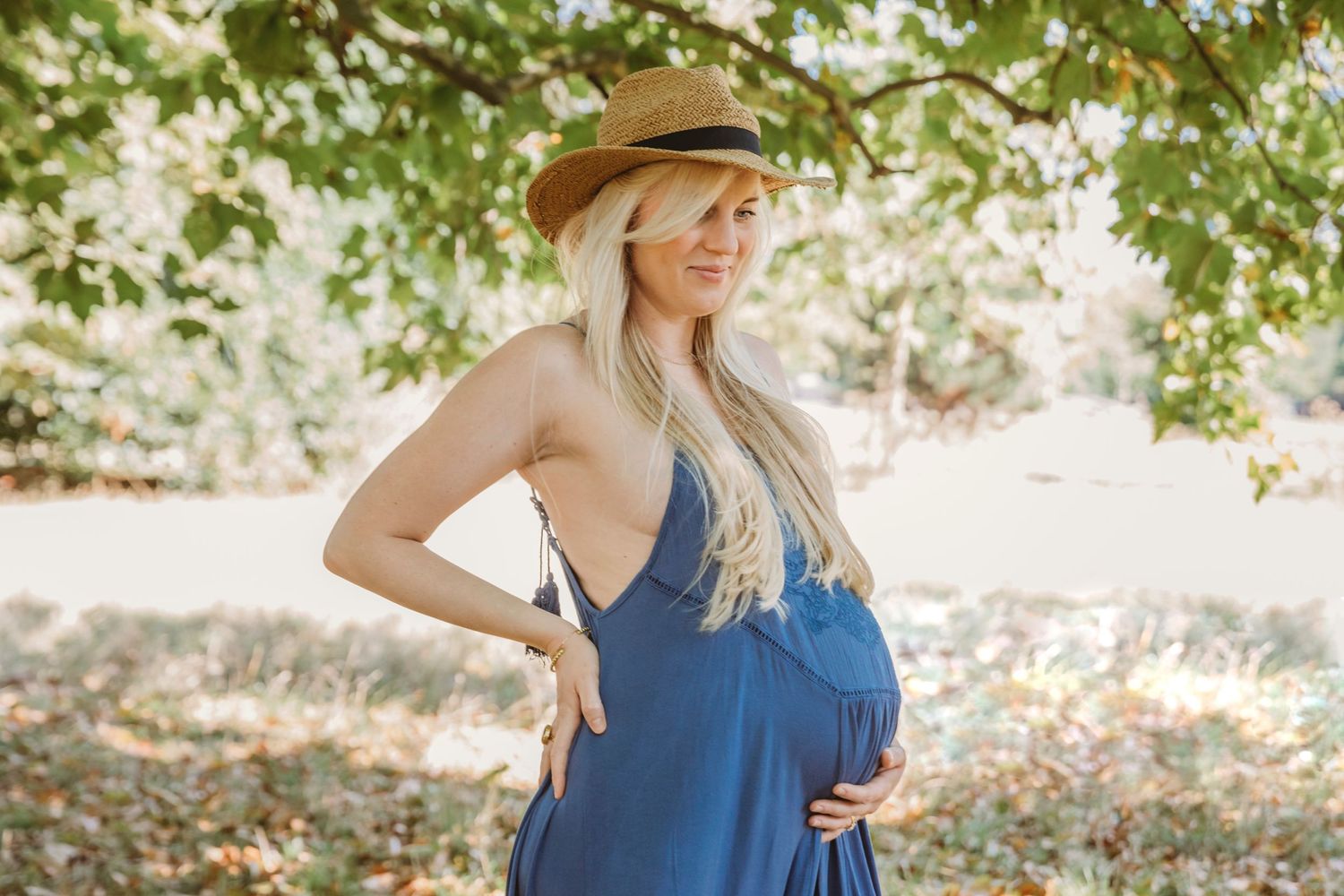 Summer maternity photo featuring blue dress and straw hat in natural setting.