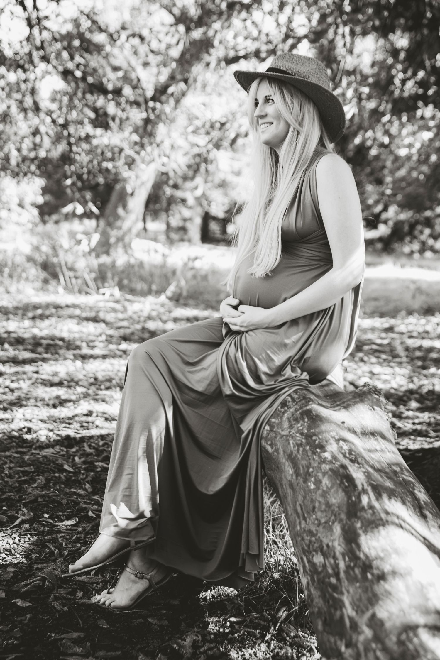 A pregnant woman in a flowing dress and hat sits on a log in a forest setting.