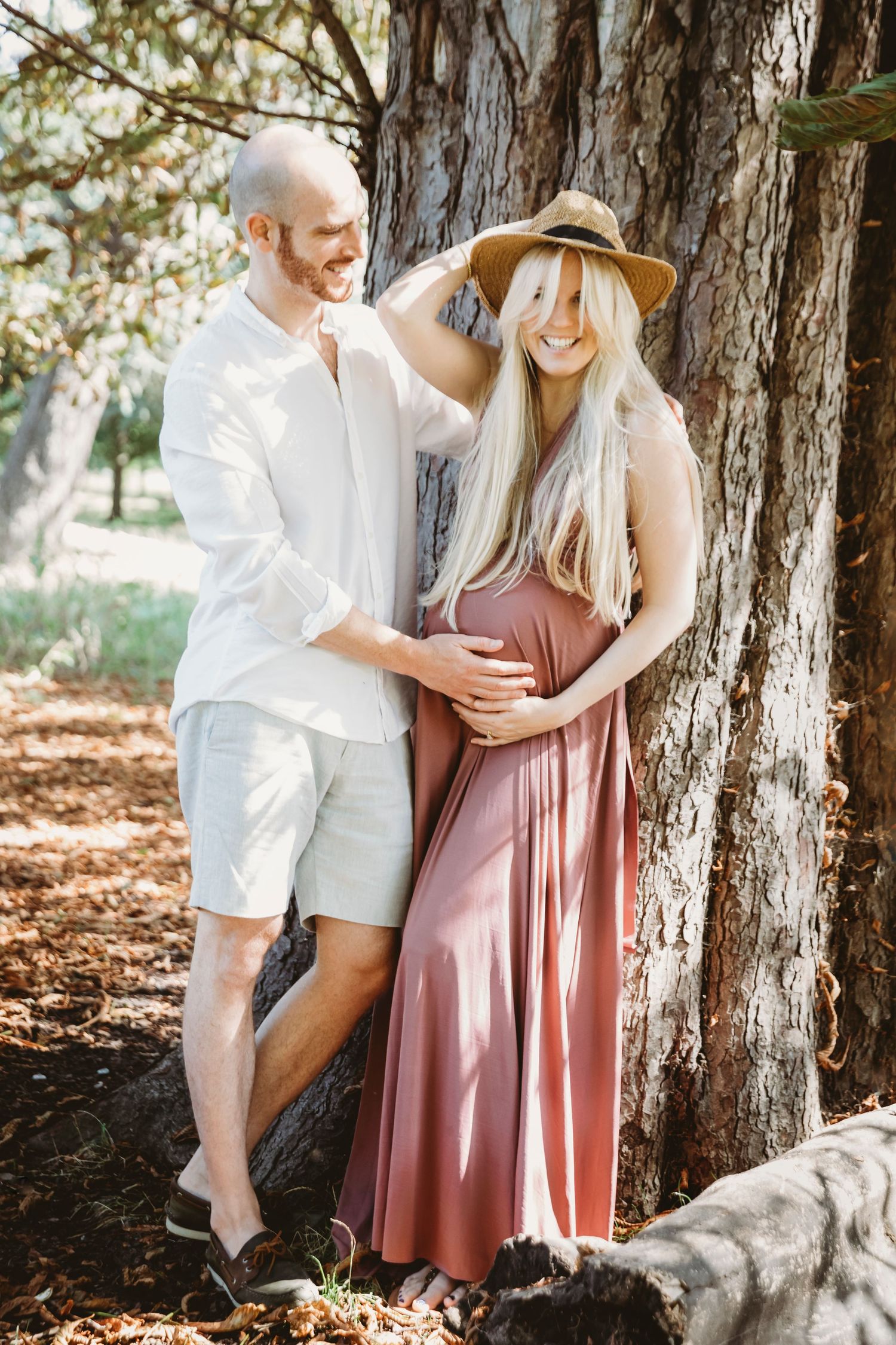 A tender maternity moment captured between a couple leaning against a large tree.