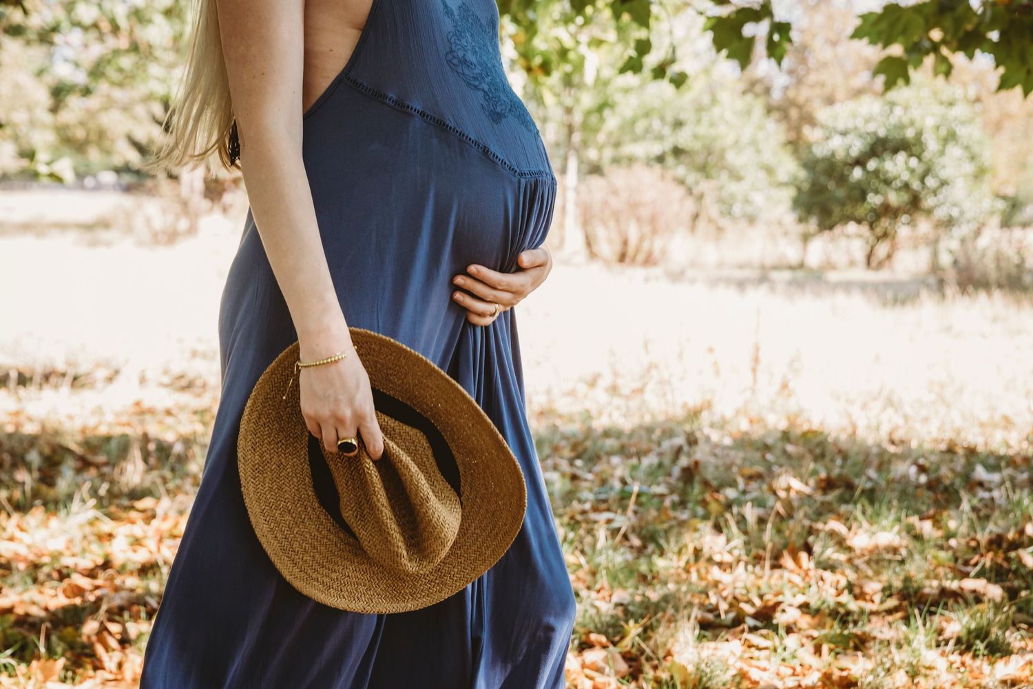 Person in blue dress holding straw hat while showing baby bump in outdoor setting.