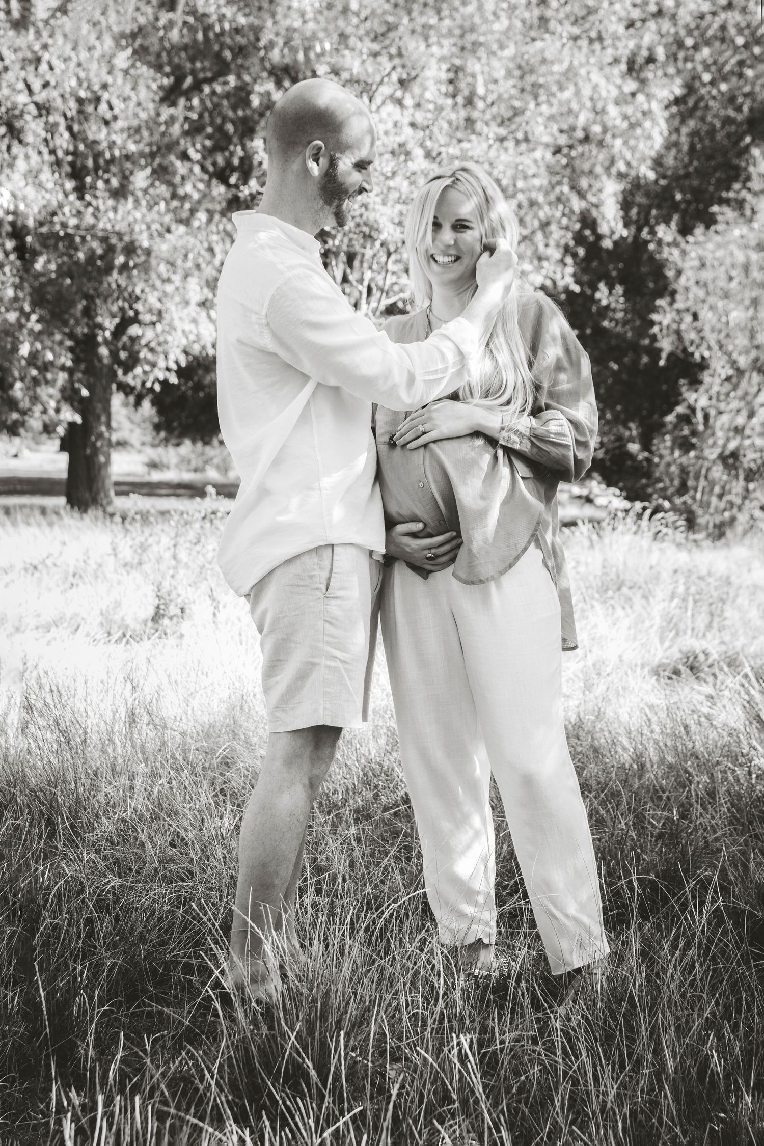 Expecting couple embracing outdoors in a sunny field wearing casual white clothing.