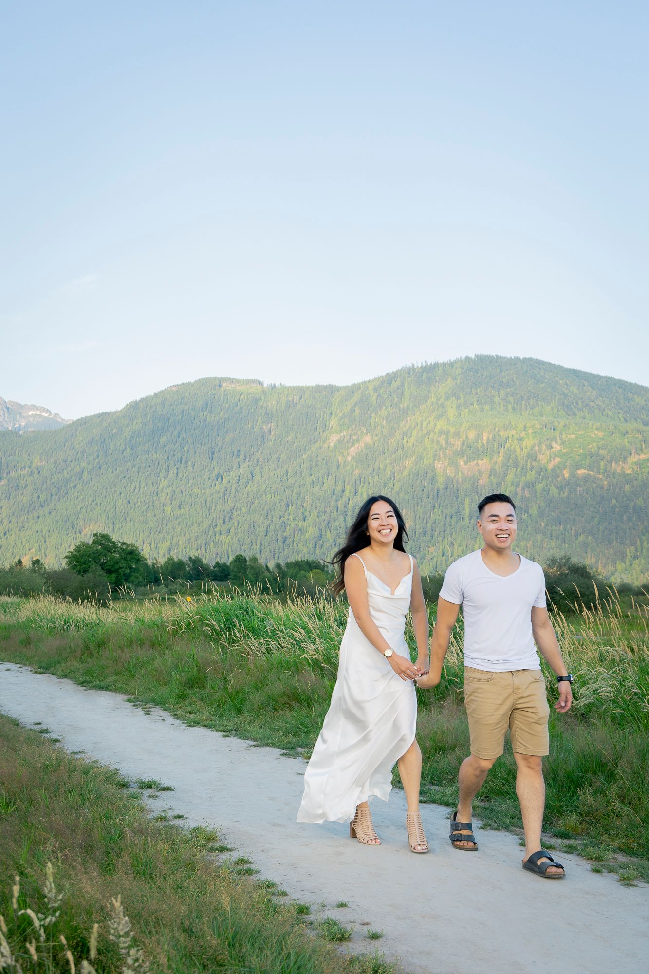 A man and woman walk hand in hand towards camera, laughing and celebrating their engagement. White dress, white t-shirt.
