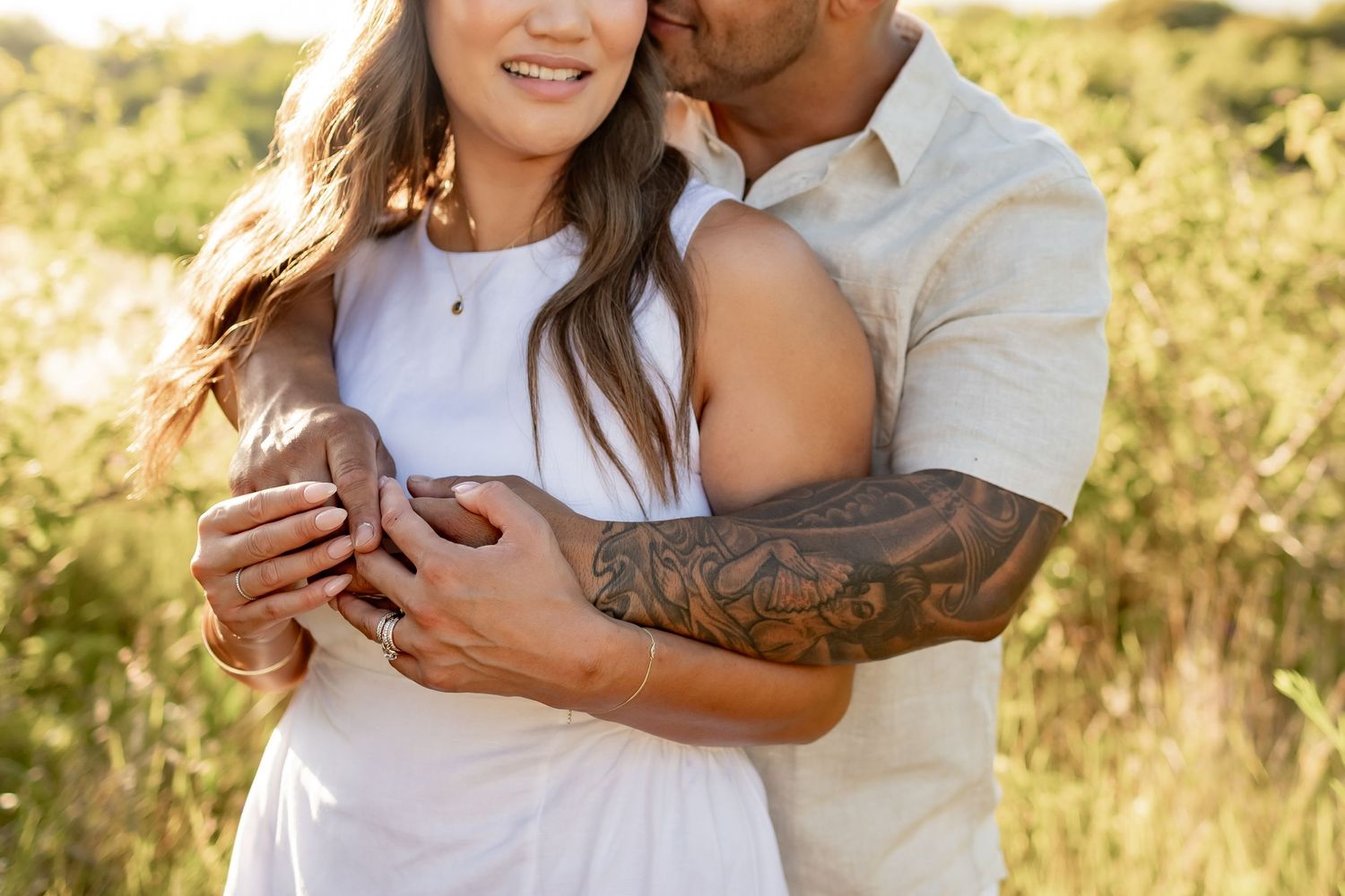 Couple embraces in a sunlit field with tall grass while dressed in white casual clothing.
