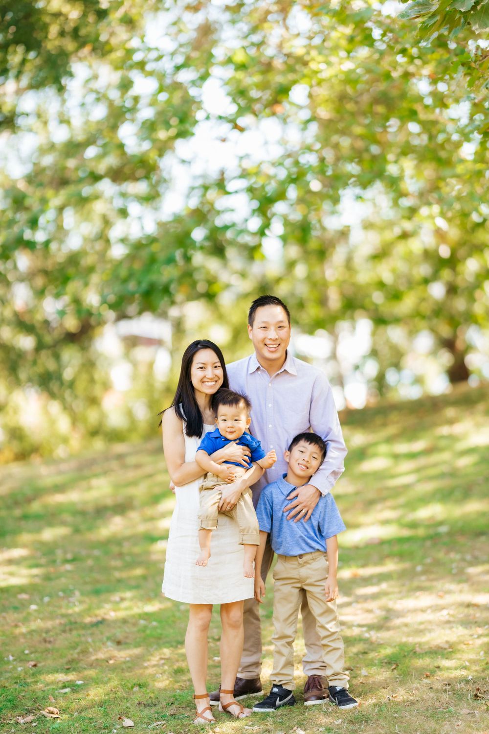 A family of four poses together on a grassy hillside under leafy trees.