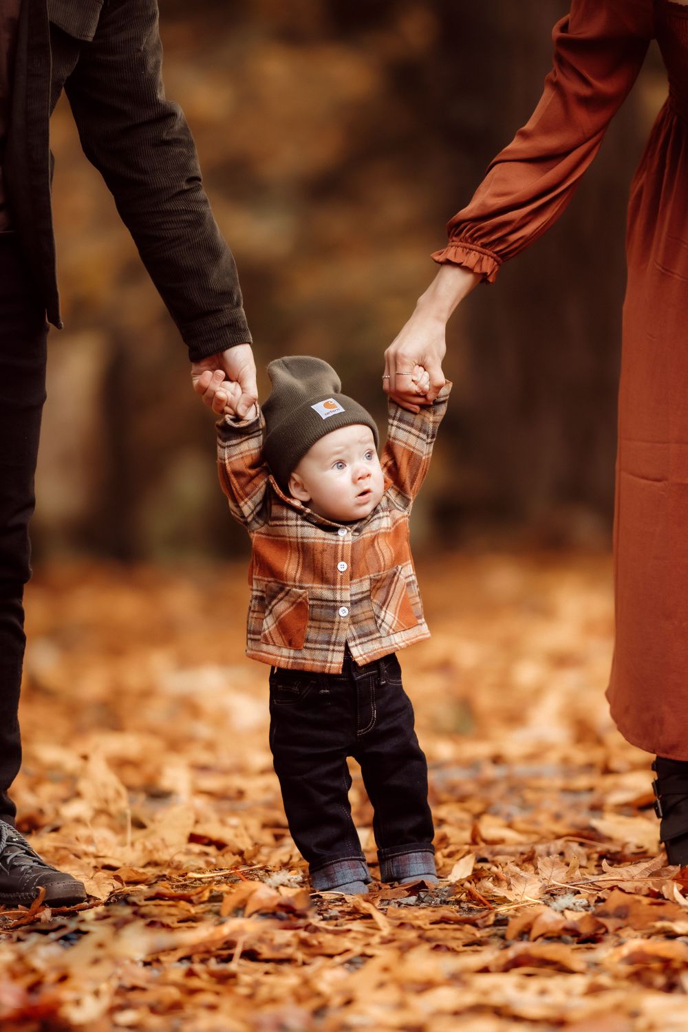 Small child in plaid shirt holding parents' hands while standing in fallen leaves.