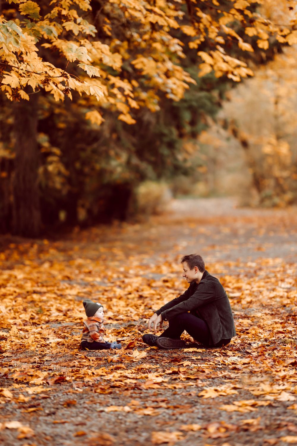 Parent and child sitting together on path covered in golden autumn leaves.