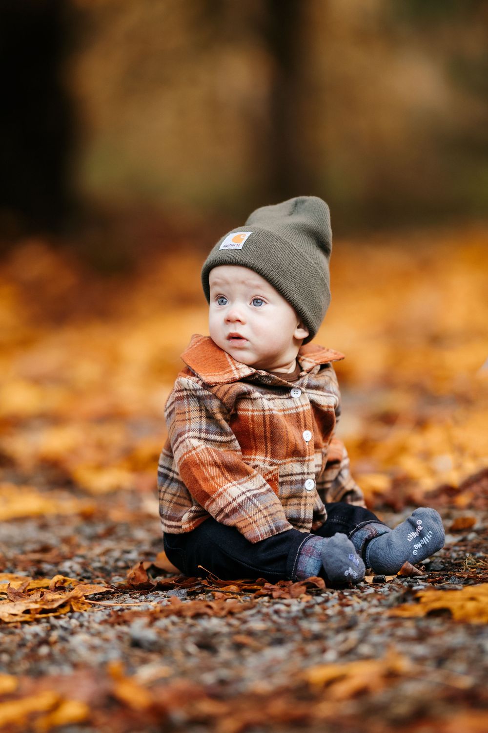 Baby wearing plaid shirt and beanie sits among fallen autumn leaves.