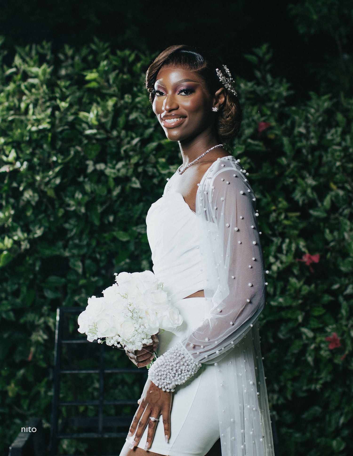 A bridal portrait with pearl-detailed sleeves and white bouquet against green foliage.