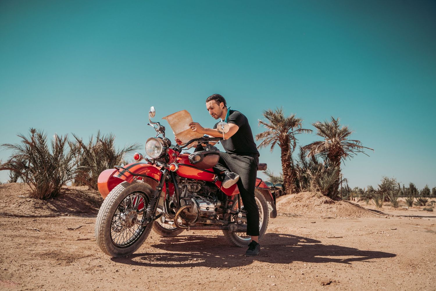 A vintage red motorcycle being ridden through a desert landscape with palm trees.