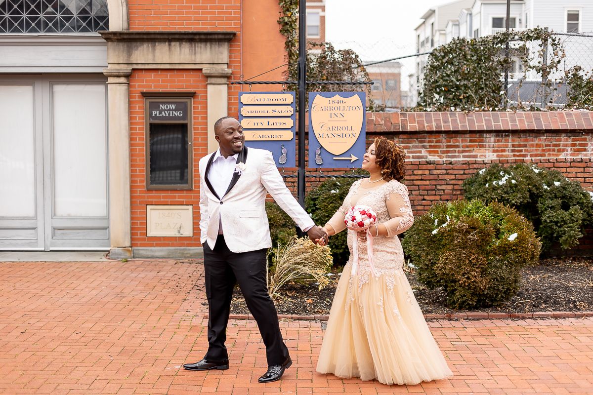 Couple in wedding attire holding hands on brick path, groom in white tuxedo, bride in gold gown.