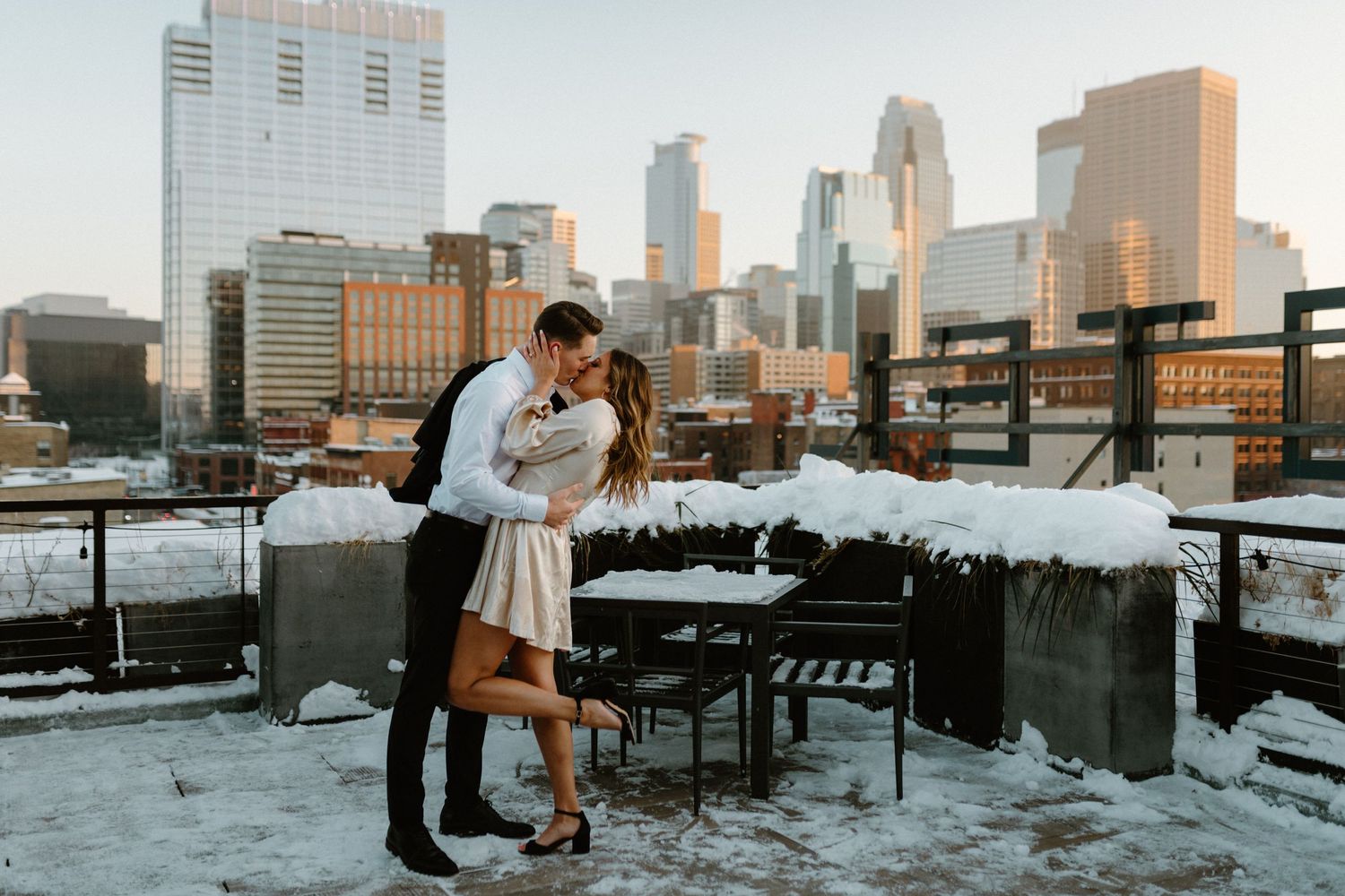 Couple embracing on snow-covered rooftop with city skyline in background during winter.