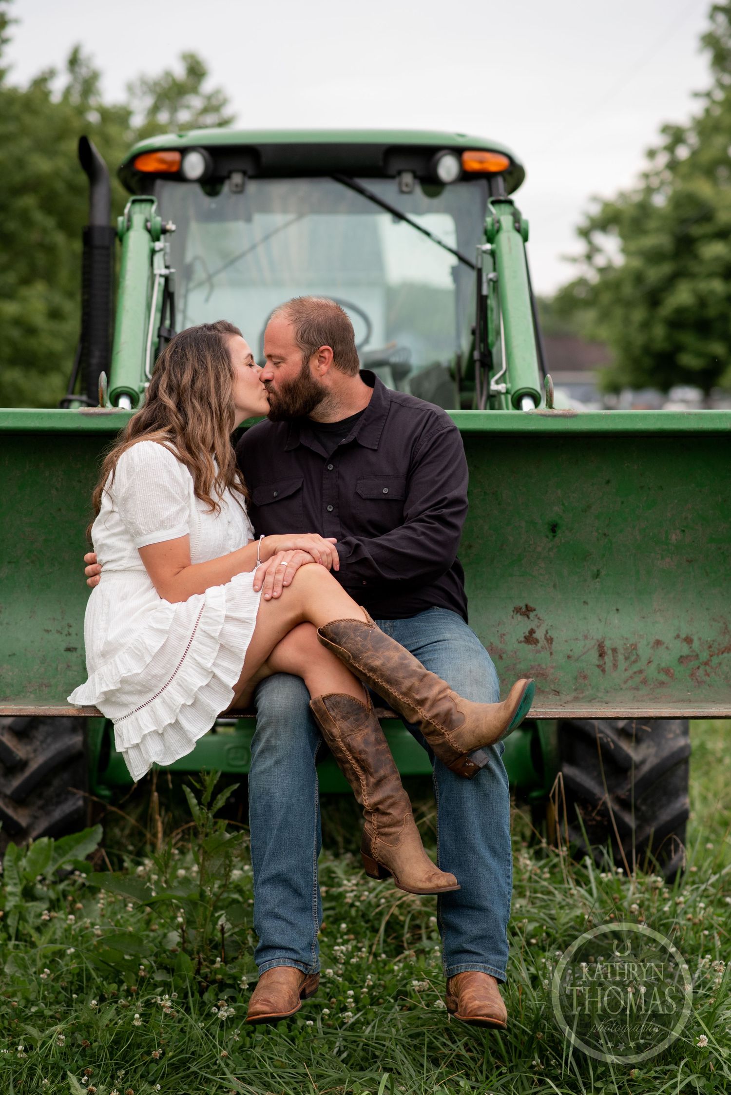 A couple embraces while sitting on the front of a green tractor in a pastoral setting.