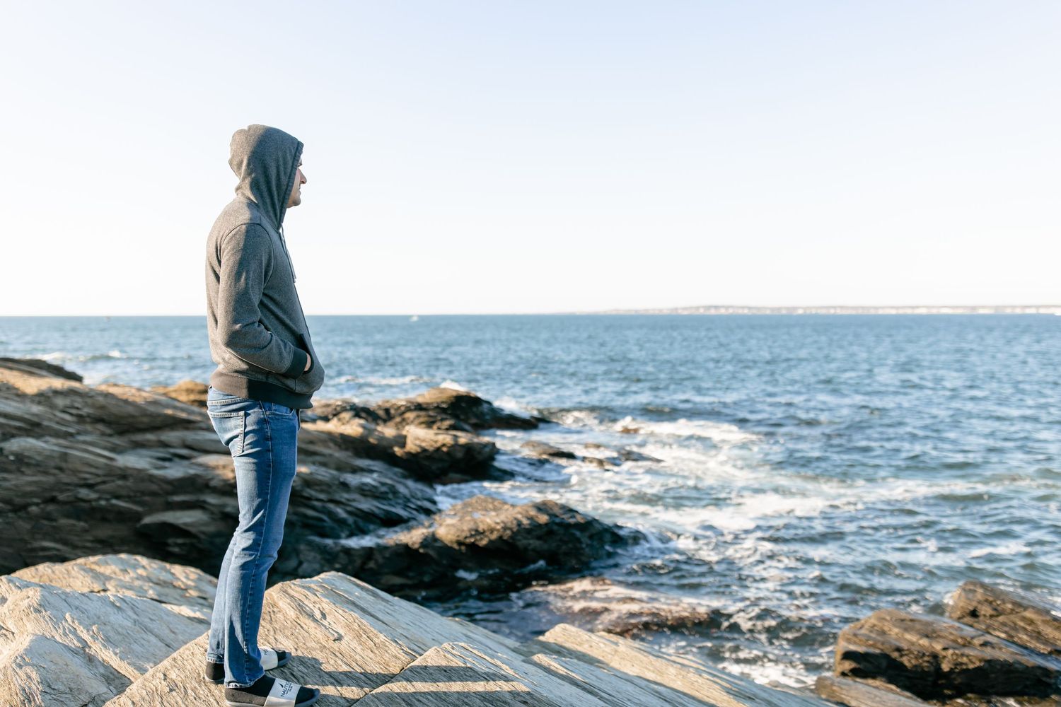 Person in gray hoodie standing on rocky New England coastline gazing at the ocean.