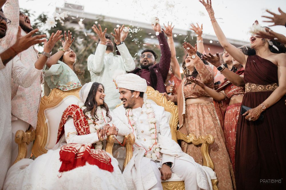 Joyous wedding celebration with guests throwing flowers and cheering while couple sits in traditional Indian attire.